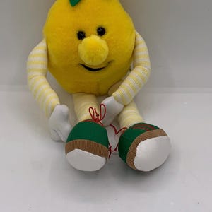 May include: A plush yellow lemon character with a smiling face, black eyes, and a small nose. It has striped arms and legs, white hands, and green and white feet. A gold halo sits atop its head. The toy is sitting down.