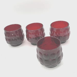 May include: A set of four small, deep red glass tumblers featuring a rounded, bulbous shape with a textured, raised circular pattern across the exterior surface. Each glass has a wide, open top and a smooth, circular rim, showcasing a consistent ruby color.