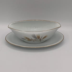 May include: White porcelain gravy boat and saucer set. The gravy boat has a curved shape with a spout and a gold rim. The saucer is round with a gold rim. Both pieces feature a wheat stalk design in gold and gray.