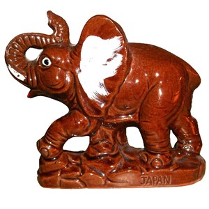 May include: A brown ceramic elephant figurine with a white ear, standing on a brown base. The base has the word "JAPAN" inscribed on it.
