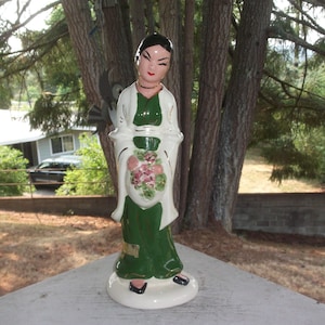 Puede incluir: Una figurita de cerámica de una mujer con un vestido verde y una chal blanca, sosteniendo un ramo de flores rosas. La figurita está de pie sobre una base blanca.