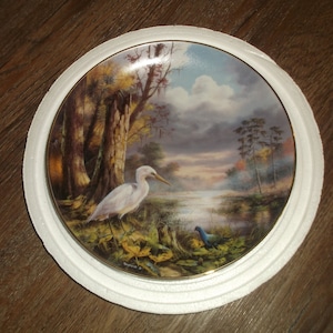 May include: A white porcelain plate with a gold rim features a painting of a white egret standing in a swamp with a blue bird in the foreground. The background is a misty, colorful landscape with trees and a river.
