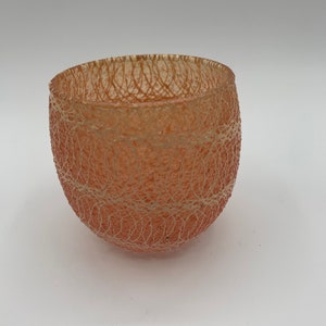May include: A peach-colored, round glass bowl with a textured, web-like pattern. The bowl has a wide opening and a slightly irregular shape. The intricate design gives the bowl a delicate appearance. The bowl is likely used for decorative purposes.