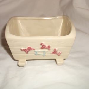 Könnte beinhalten: Ein weißer Keramik-Pflanztopf in Form einer Babywiege mit rosa und blauen Akzenten. Der Topf hat vier Beine und ist mit drei rosa und blauen Schleifen verziert.