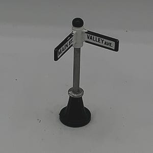 May include: A miniature street sign with a black base and a silver pole. The sign has two arms, one reading "MAIN ST." and the other "VALLEY AVE." in white text. The top of the pole has a black sphere.