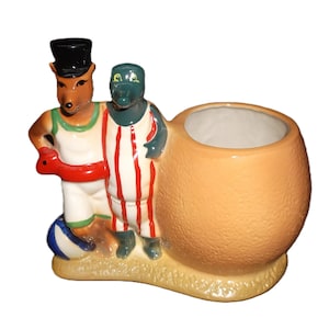 May include: A ceramic planter shaped like an orange with a red and white striped alligator and a brown fox wearing a top hat and holding a red and white striped life preserver.