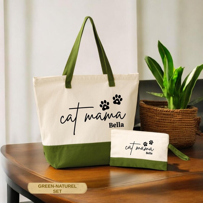 A green and white tote bag with the text "cat mama Bella" printed on it, along with paw prints, sits on a wooden surface next to a small woven basket and a potted plant.