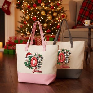 May include: Two off-white canvas tote bags with pink and gray bottom panels. The front of each bag features the word JOY in festive patterns, a Santa hat, holly leaves, and the name Jessica printed in red script below the graphic design.