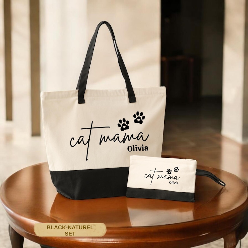 A white tote bag with black trim and paw prints, and a smaller white bag with the same paw print design, both labeled "cat mama Olivia" on a wooden table.