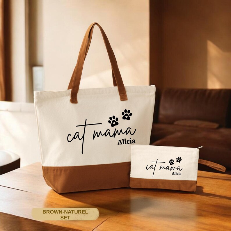 A tote bag with the text "cat mama Alicia" printed on it, along with paw prints, sits on a wooden surface. There is also a smaller bag with the same text and paw prints.