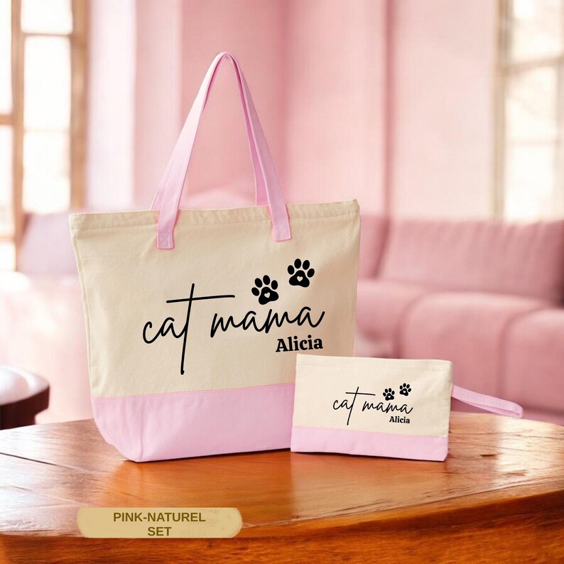 A pink tote bag with the text "cat mama Alicia" printed on it, along with paw prints, sits on a wooden surface. Next to the bag is a smaller white bag with the same text "cat mama Alicia" printed on it. The background is a pink color, possibly a couch or chair.