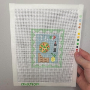 May include: Needlepoint canvas featuring an Italian theme. The design includes a pizza, wine bottle, and lemon, with the word "ITA" and an Italian flag. The canvas is white with a green border and the text "KTNDLPTCO".