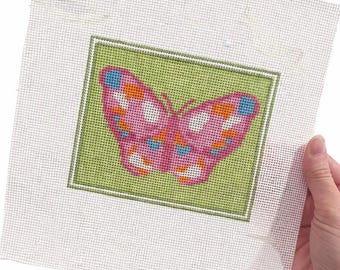 May include: A needlepoint canvas featuring a pink butterfly design with orange, blue, and white accents. The butterfly is set against a green background within a white border. The canvas is held by a hand.