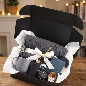 May include: A black gift box lined with white tissue paper containing a dark gray diamond-patterned knit blanket tied with a white ribbon, a gold spoon, a gray ribbed mug, a blue notebook, a black candle, and a small brown crocheted creature.