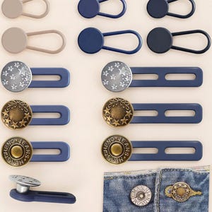 Könnte beinhalten: Auswahl an Jeansknopfverlängerungen und Taillenverstellern in verschiedenen Farben und Designs. Enthält silberne Sternenmuster, goldene Sterne und gravierte Knopfstile. Gezeigt werden auch Knopfverlängerungen mit Schlaufenverschlüssen.