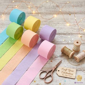 May include: A collection of six pastel crepe paper rolls in blue, yellow, green, peach, purple, and pink. Included are two wooden spools with metallic twine, a pair of copper-toned scissors, and a tag with the text Craft Your Celebration.