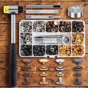 May include: A metal hammer with a yellow plastic head, a clear plastic organizer box filled with various metal snap fasteners in silver, gold, bronze, and black finishes, along with several metal setting tools and a circular metal base.