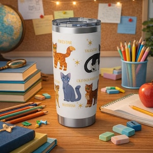 May include: A white stainless steel tumbler featuring illustrations of cartoon cats with names like Firestar and Tallstar. The tumbler has a silver lid and is set on a wooden desk with school supplies.