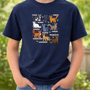 以下が含まれることがあります： 「Firestar」や「Bluestar」などの名前が付けられた猫のイラストが描かれた、ネイビーブルーのTシャツ。デザインには星が含まれています。Tシャツは人が着用しています。Tシャツは半袖です。