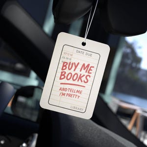 May include: A cream-colored tag with the text "BUY ME BOOKS AND TELL ME I'M PRETTY" in red, hanging from a string. The tag has a library card design with dates and the word "LIBRARY" at the bottom.