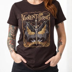 May include: Brown t-shirt with a graphic design featuring a sword, bat wings, and the text "little Violent Thing." The phrase "I will not die today" is also included. The shirt is a dark brown color.