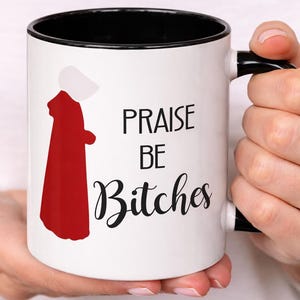 May include: White ceramic mug with a black interior and handle. The mug features the text "PRAISE BE Bitches" in black, along with a red and white graphic of a figure in a red cloak and white bonnet.