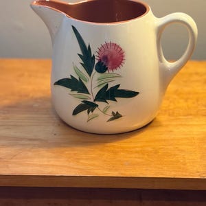 May include: White ceramic pitcher with a handle and spout, featuring a hand-painted thistle design in shades of green, pink, and purple. The interior rim is brown. The pitcher is sitting on a wooden surface.