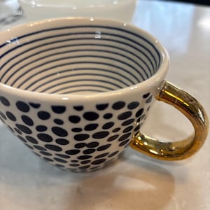 Könnte beinhalten: Weiße Keramik-Teetasse mit goldenem Henkel und marineblauem Punktmuster auf der unteren Hälfte. Die obere Hälfte hat marineblaue horizontale Streifen. Eine weiße Schüssel ist im Hintergrund leicht unscharf.