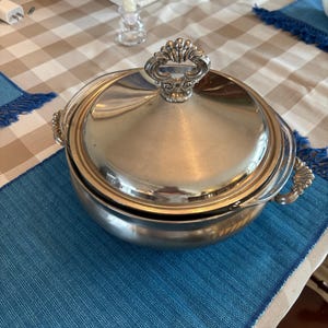 May include: A silver-colored serving dish with a lid and ornate handle. The dish has a rounded shape and sits on a blue placemat. The handle is detailed with a decorative design. The background includes a checkered tablecloth and other table settings.