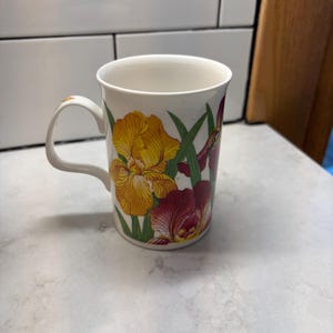 May include: A white ceramic mug featuring a botanical illustration of vibrant yellow and deep maroon iris flowers with green leaves. The mug has a smooth, cylindrical body and a curved handle, showcasing detailed floral patterns on the exterior surface.
