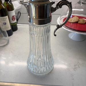 May include: A clear glass carafe featuring a vertical ribbed pattern and a flared base. The vessel is topped with a polished silver-toned metal lid and a matching ornate, curved handle, creating a distinct contrast between the glass and metal.