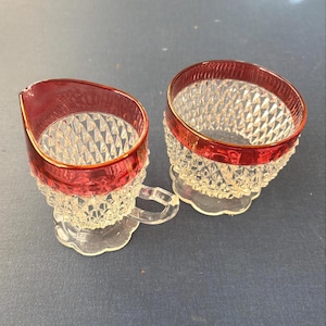 May include: Two vintage clear glass cream and sugar set. The rims are a vibrant red with a gold trim. The glass has a diamond pattern. The creamer has a handle and a spout. The sugar bowl has a matching red rim.