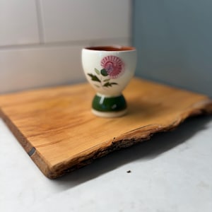 May include: A white ceramic egg cup with a pink and purple floral design. The egg cup has a green base and a brown interior. It is sitting on a wooden cutting board.