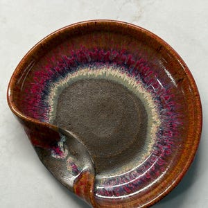May include: A round ceramic spoon rest featuring a dark brown center and a wide, textured rim with a reactive glaze in shades of deep red, magenta, and blue. The edge of the dish curves inward on one side to create a functional indentation.