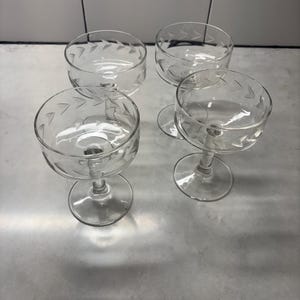 Könnte beinhalten: Vier klare Champagner-Coupe-Gläser. Jedes Glas hat eine runde Schale mit einem dekorativen geätzten Muster und einem Stiel. Die Gläser sind auf einer hellen Oberfläche angeordnet.