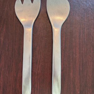 May include: A pair of silver-colored salad servers. One server is a fork with three tines, and the other is a spoon-shaped server. Both have long, rectangular handles. The servers are displayed on a dark wood surface.