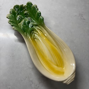 May include: A ceramic serving dish shaped like a stylized head of lettuce. The dish features a green, textured top representing the leaves, transitioning to a creamy white and yellow interior. The dish is designed to resemble a halved lettuce.