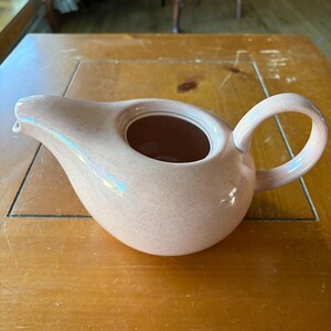 May include: A light pink ceramic gravy boat with a speckled finish, featuring a wide pouring spout, a rounded body, and a curved handle on the side. The interior of the vessel is smooth and hollow, designed for pouring liquids or sauces.