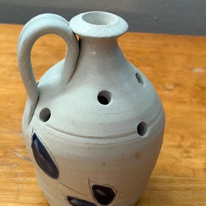 May include: A light blue ceramic pitcher with a handle and a round opening at the top. The pitcher has several holes around the body and is decorated with dark blue leaf-like designs. The pitcher is sitting on a wooden surface.