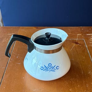 May include: A white teapot with a black handle and a black lid. The body of the teapot features a blue floral pattern consisting of small flowers and leaves. A metallic silver band encircles the upper portion of the teapot just below the lid.