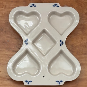 May include: Off-white ceramic baking dish with heart-shaped and diamond-shaped compartments. The dish has a unique hourglass shape with blue floral accents. A charming kitchenware item for baking or serving.