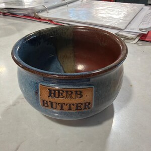 May include: A round ceramic crock with a blue and brown glazed finish. The exterior features a rectangular embossed plaque with the text HERB BUTTER in dark lettering. The interior is divided into two distinct color sections of blue and brown.