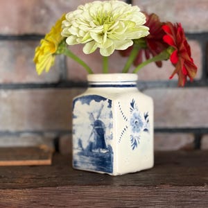 May include: A square ceramic vase with a cream-colored base and blue painted details. One side features a blue windmill landscape, while another side displays a blue floral pattern. The vase holds three flowers: one pale yellow, one bright yellow, and one red.