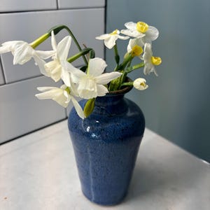 May include: A small, blue ceramic vase holding a bouquet of white daffodils with yellow centers. The vase has a textured surface and a dark rim. The flowers are in full bloom, with green stems and leaves.