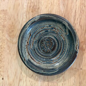 May include: A handmade ceramic dish with a blue and brown glaze. The small, round dish features a spiral design and a central embossed image. The dish is approximately 4 inches in diameter and is suitable for holding small items.