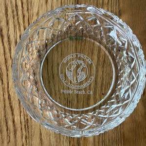 May include: A clear, round glass bowl with a diamond-cut pattern. The center of the bowl features the text "WORLD PRO-AM GOLF TOURNAMENTS Pebble Beach, CA" in a circular design. The bowl is likely a souvenir or decorative item.
