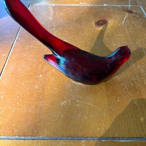 May include: A deep red glass bird figurine with a smooth, polished surface. The bird is depicted in a stylized, abstract form with a long, upward-curving tail and a rounded body, capturing light through its translucent, rich crimson glass material.