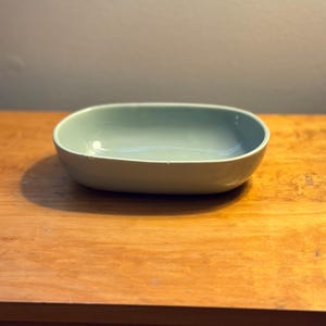 May include: A light green, oval-shaped ceramic bowl. The bowl has a smooth, glossy finish and is set on a wooden surface. The bowl's design is simple and modern, suitable for serving food or as a decorative piece.