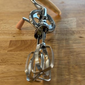 May include: A vintage hand-crank egg beater with a wooden handle and a peach-colored knob. The beater is made of shiny metal and has a gear mechanism. The blades are made of thin metal strips. The kitchen tool is on a wooden surface.