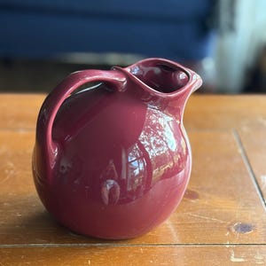 May include: A deep burgundy ceramic pitcher with a rounded, bulbous body and a smooth, glossy finish. The vessel features a curved handle on the side and a wide, integrated pouring spout at the top, showcasing a solid, uniform color throughout.